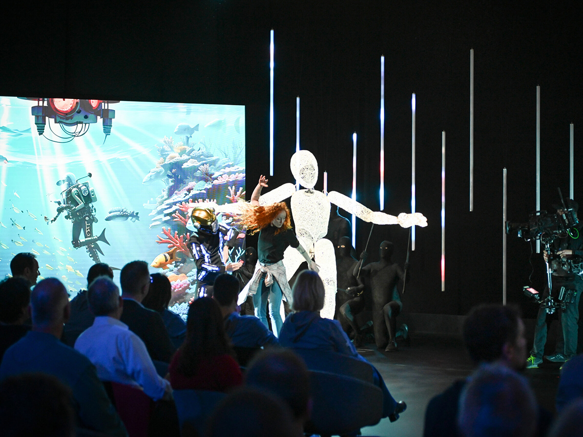 Dundu, a three-meter illuminated puppet figure, with outstretched arms behind a dancer in the foreground and a large LED screen displaying digital content.