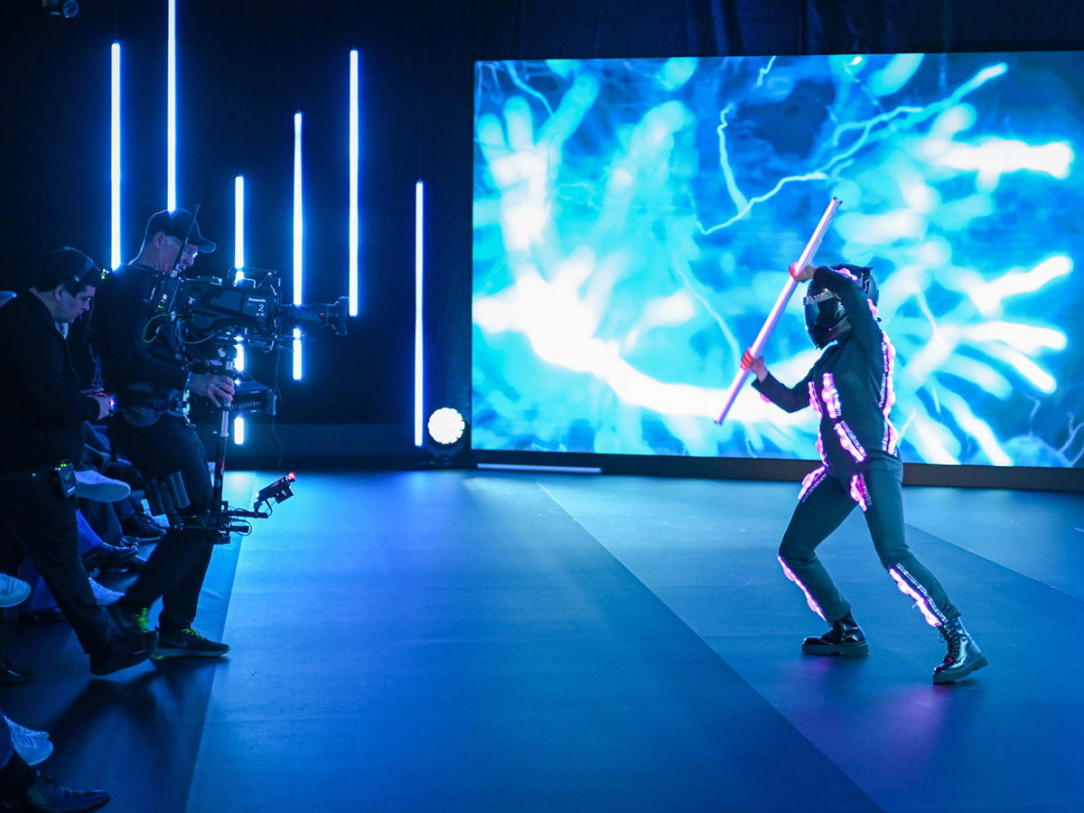 LED-suit performer on stage in front of a large LED screen showing abstract blue digital content, with camera crew filming from the side &mdash; illustrating the hybrid production setup for onsite and virtual audiences.