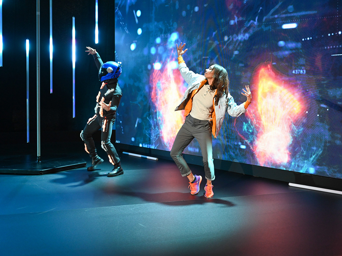 Two dancers performing in front of a large LED screen showing fiery digital content, embodying the emotional arc of the show.