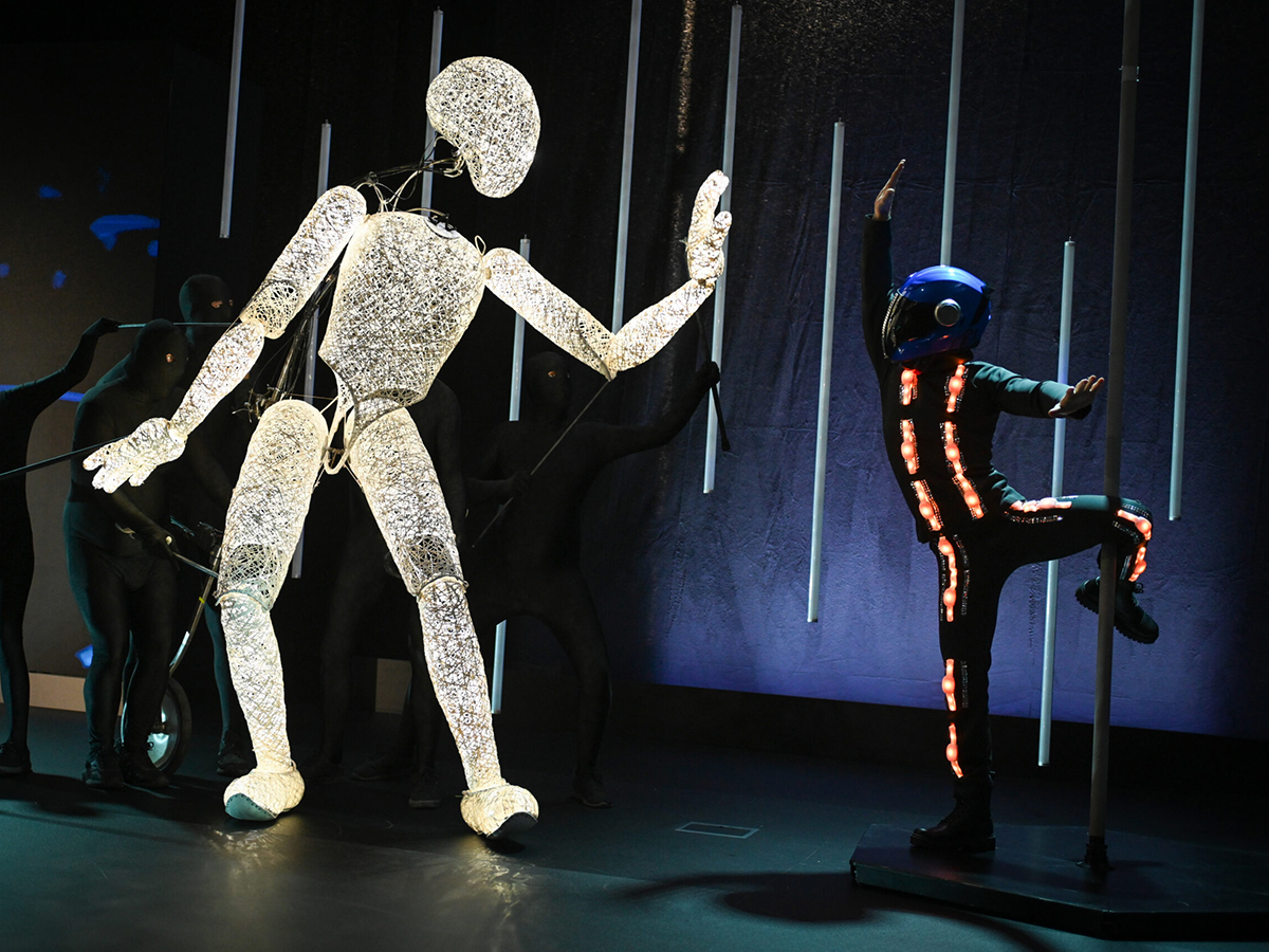 Dundu, a three-meter illuminated puppet figure, facing an LED-suit dancer on stage &mdash; two representations of AI in dialogue.