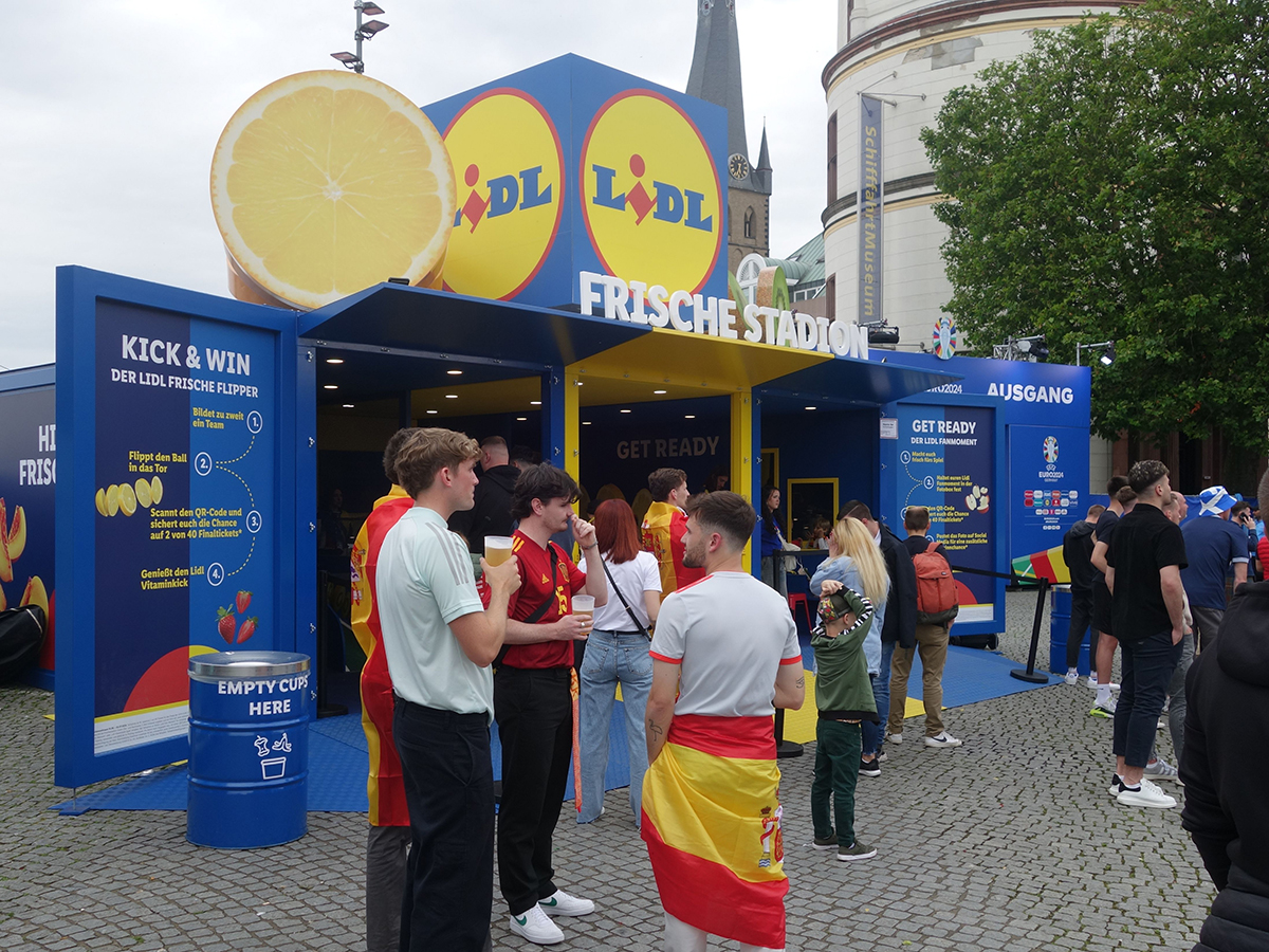 The Lidl Frische Stadion at the UEFA EURO 2024 Fan Zone in Düsseldorf, showing the branded blue container structure with oversized lemon on top and fans gathered outside.